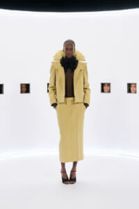 Model wearing a pale yellow tailored suit with a large black ruff collar, standing in a white studio with hands in pockets.