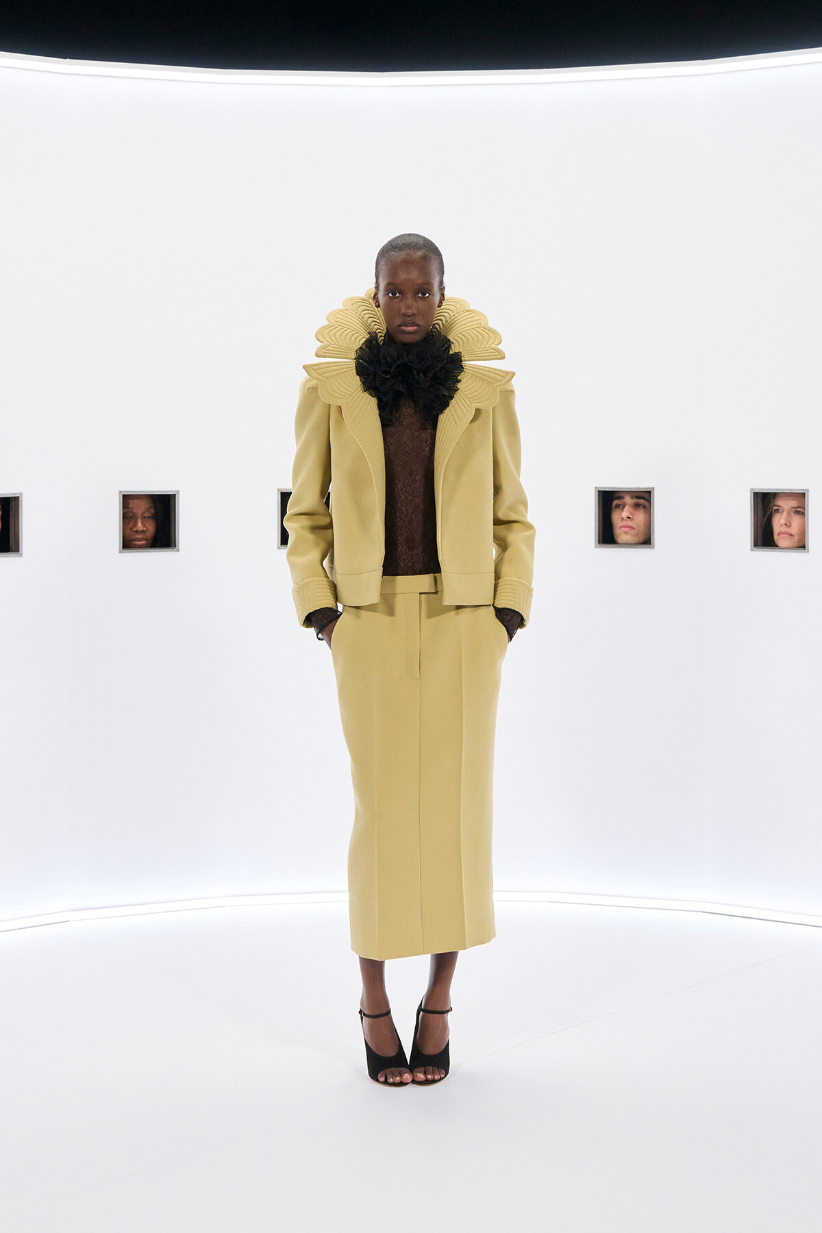 Model wearing a pale yellow tailored suit with a large black ruff collar, standing in a white studio with hands in pockets.