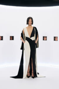 Model wearing a black and cream draped gown with a deep V neckline, standing in a white studio set.