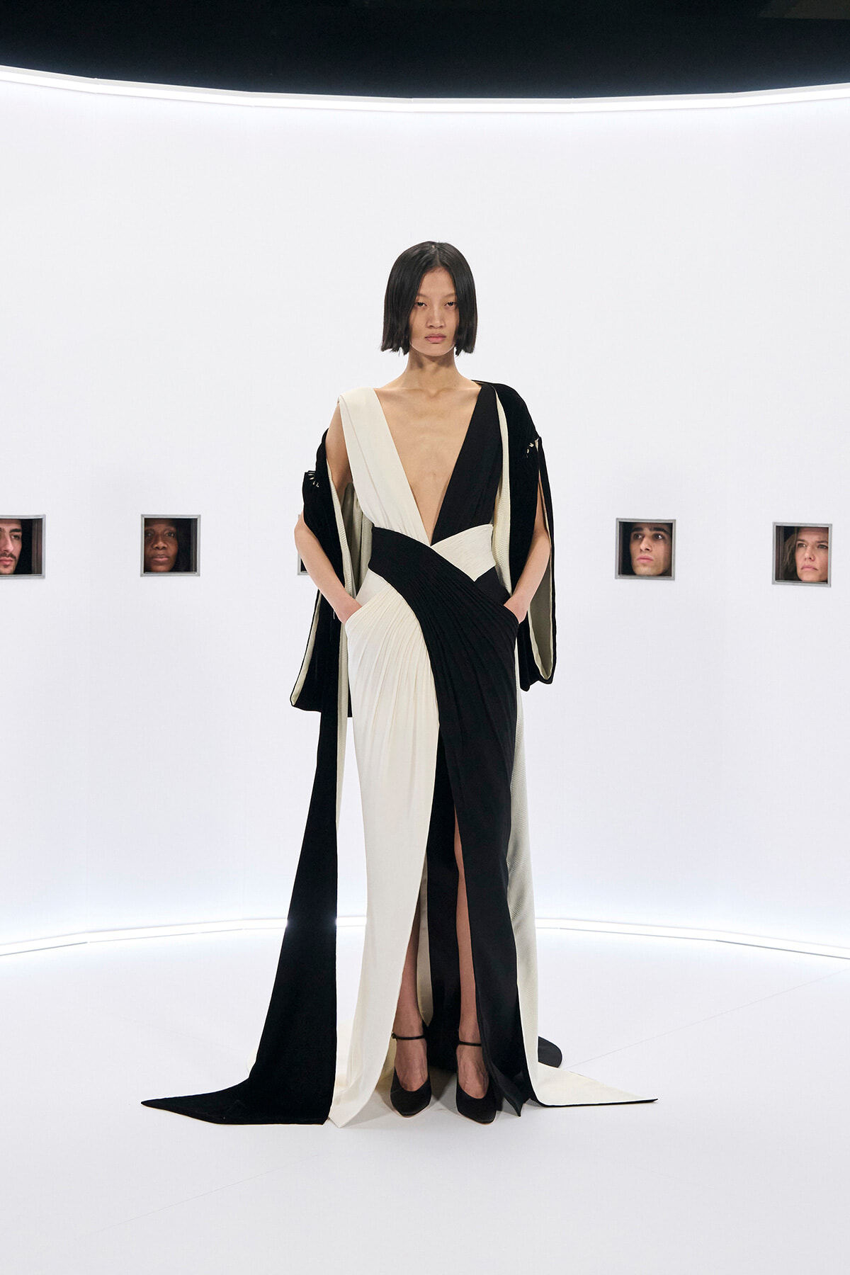 Model wearing a black and cream draped gown with a deep V neckline, standing in a white studio set.