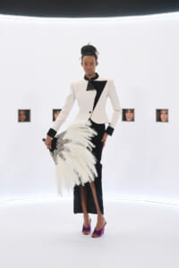 Model in a white and black tailored blazer, black skirt, and purple heels stands in a white studio holding a large white feather bouquet.