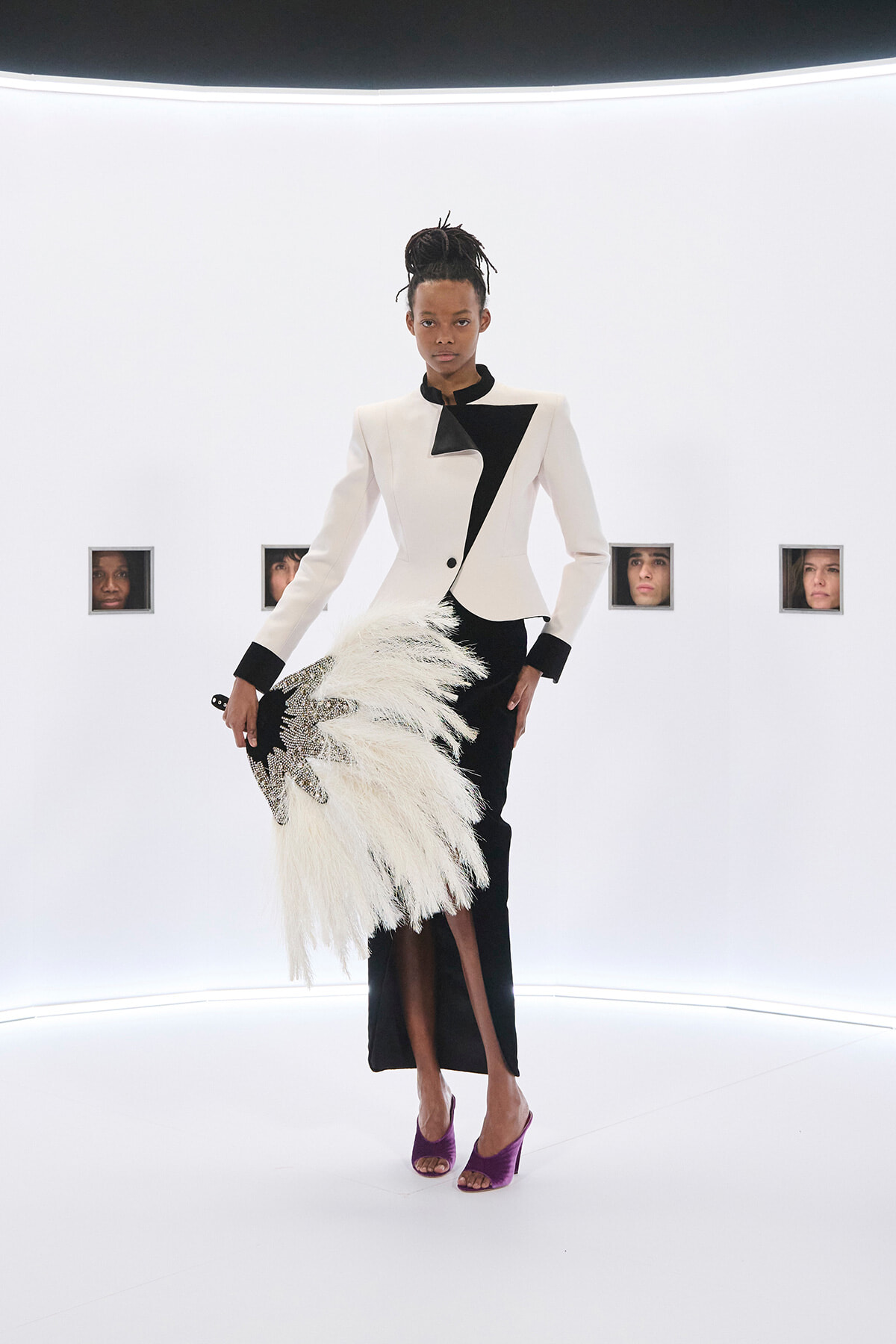 Model in a white and black tailored blazer, black skirt, and purple heels stands in a white studio holding a large white feather bouquet.