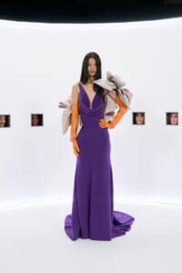 Model wearing a deep purple evening gown with long orange gloves and a large beige cape, standing in a stark white photography studio with small square windows showing observers nearby.