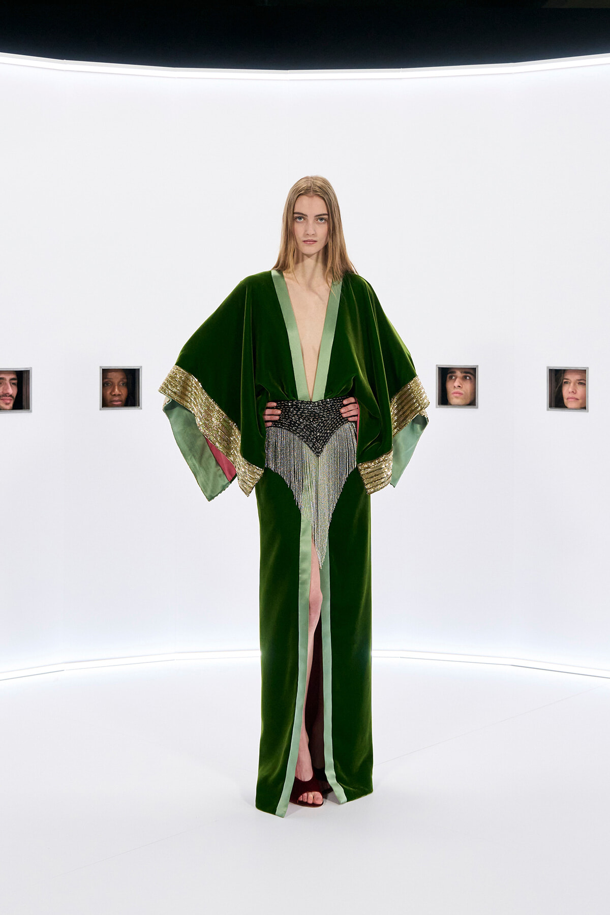 Model on a white studio runway wearing a dark green velvet gown with light green trim, gold-embellished cuffs, and a beaded fringe belt; hands on hips.
