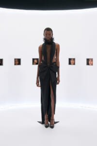 Model wearing a black evening gown with a sheer plunging bodice, large waist bow, and high slit, standing in a white gallery with small framed faces on the wall behind.