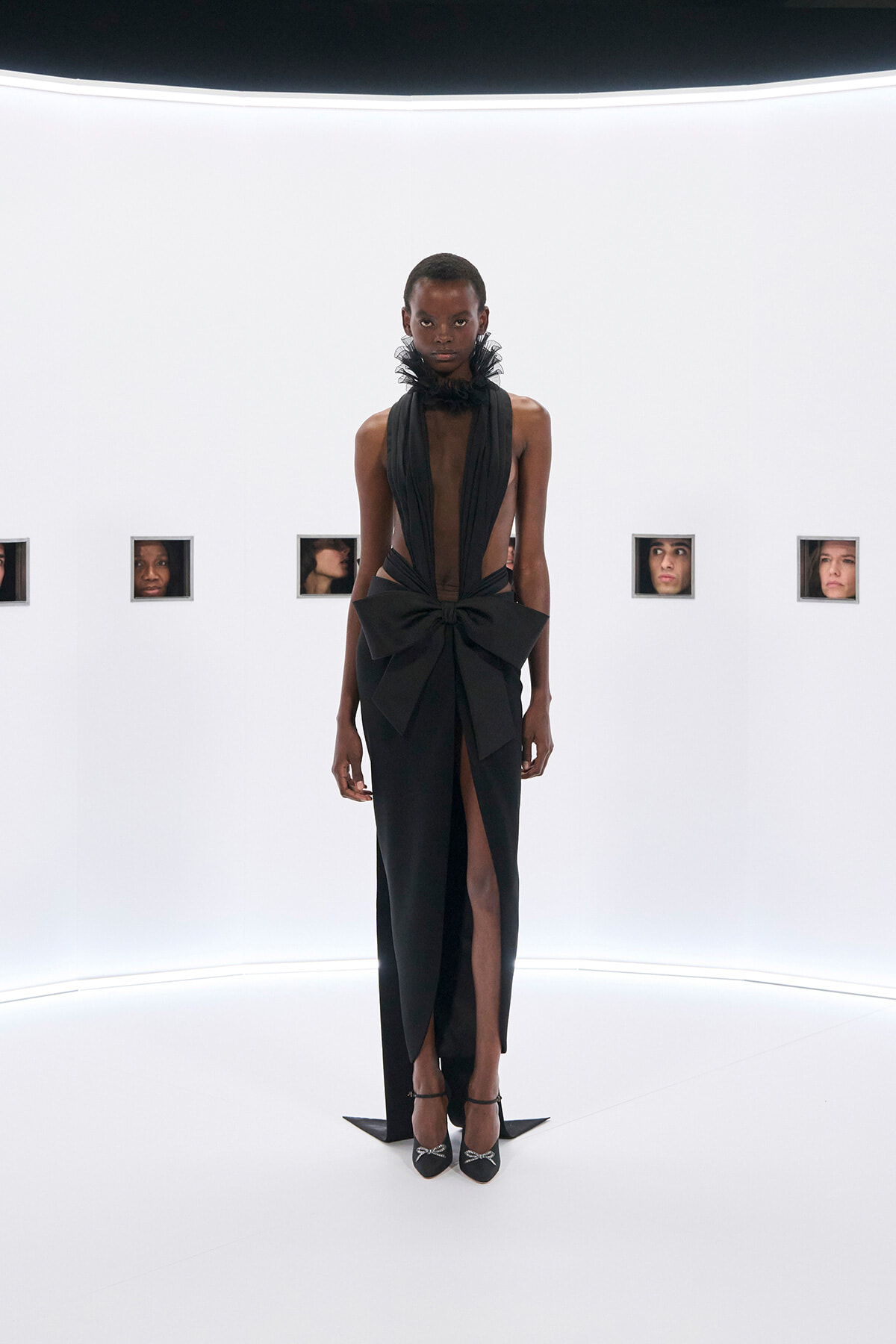 Model wearing a black evening gown with a sheer plunging bodice, large waist bow, and high slit, standing in a white gallery with small framed faces on the wall behind.