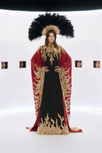 Model in a red and gold flame-embellished cape over a black dress, wearing a large black feather headdress in a white studio setting.