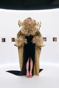 Model in a gold sequined gown with a large, ruffled collar and sleeves, standing in a white gallery with small framed portraits in the background.