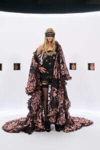 Model wearing a black and pink lace cape and fitted outfit, with a black masquerade mask, standing in a stark white studio with small window openings behind her.