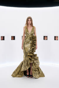 Model wearing a gold metallic gown with a plunging V-neck and dramatic pleated ruffle skirt, standing in a white studio.