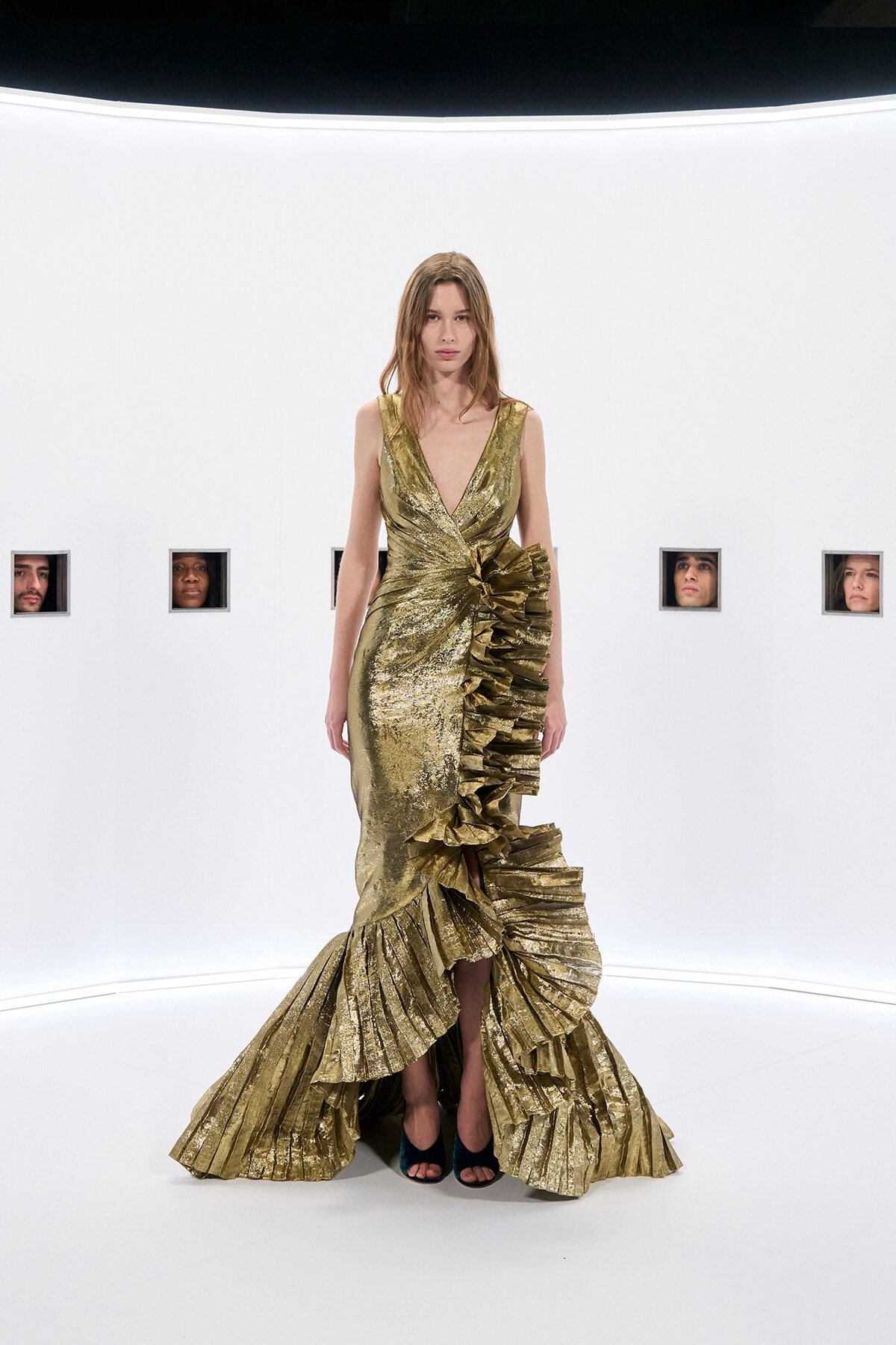 Model wearing a gold metallic gown with a plunging V-neck and dramatic pleated ruffle skirt, standing in a white studio.