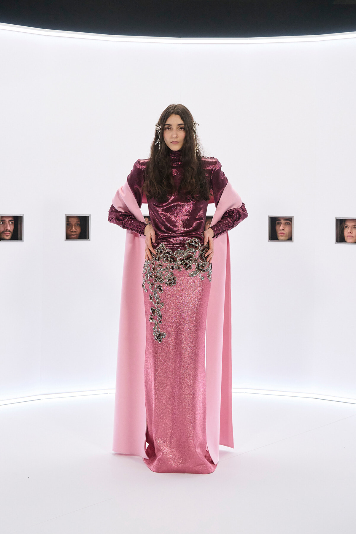 Center-stage model wearing a metallic pink gown with a matching pink cape, hands on hips in a bright white studio setting.