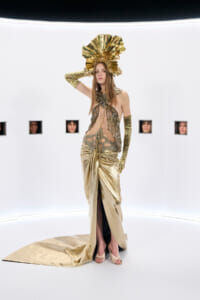Model in a gold metallic gown with an ornate headdress, posing in a stark white exhibition space with small wall openings showing faces behind.