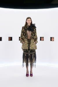 Model in a gold-embellished black coat over a sheer black dress with a large black bow, standing in a white studio backdrop.
