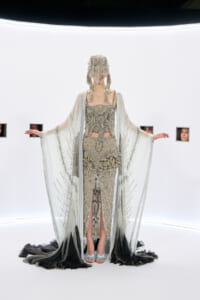 Model in an ornate silver beaded gown with a matching cape and fringe headpiece in a stark white gallery setting.