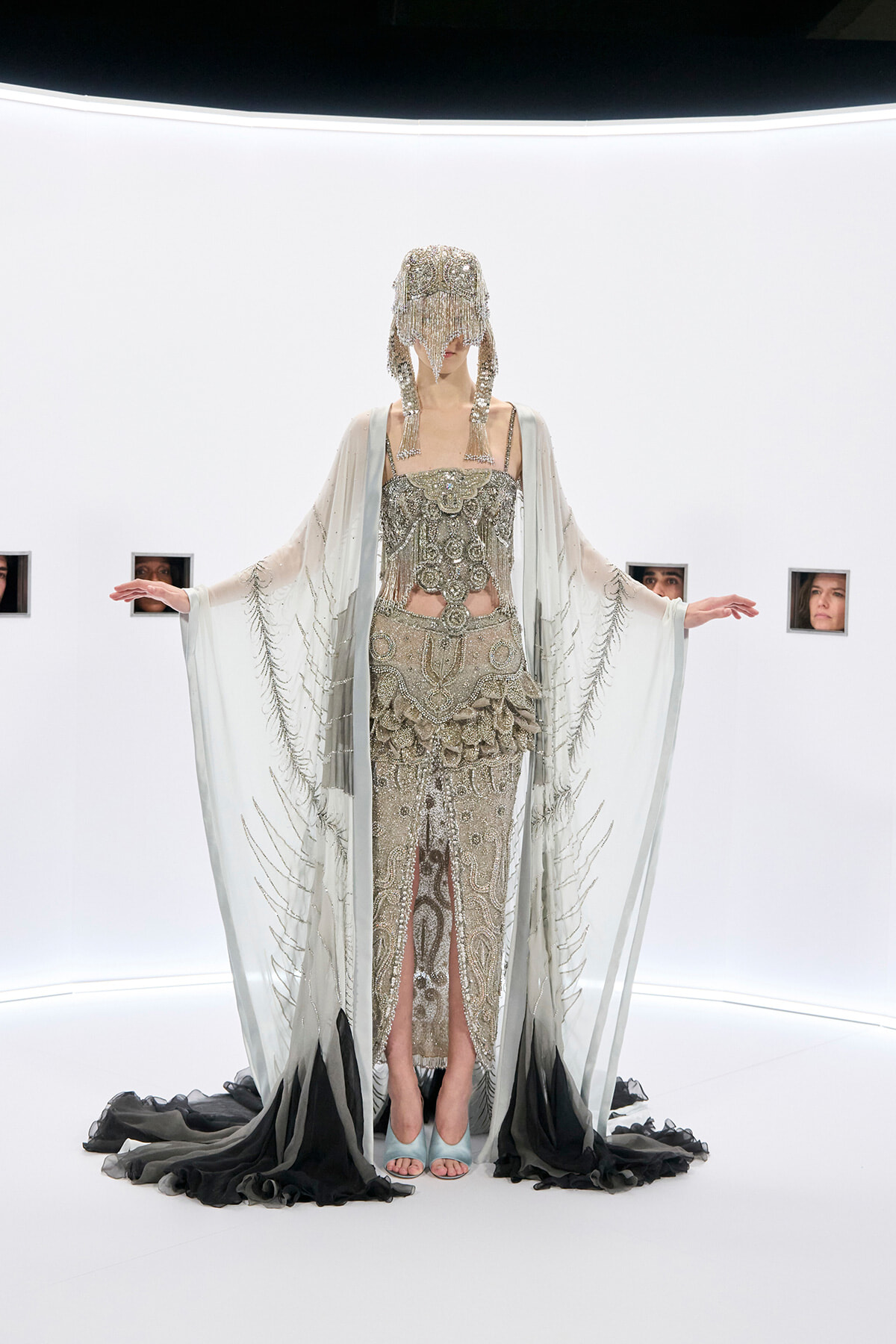 Model in an ornate silver beaded gown with a matching cape and fringe headpiece in a stark white gallery setting.