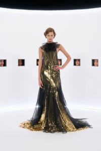 Model in a full-length gold sequin gown with black sheer overlay and a large bow at the neck, posing in a stark white studio with small square window cutouts showing faces in the background.