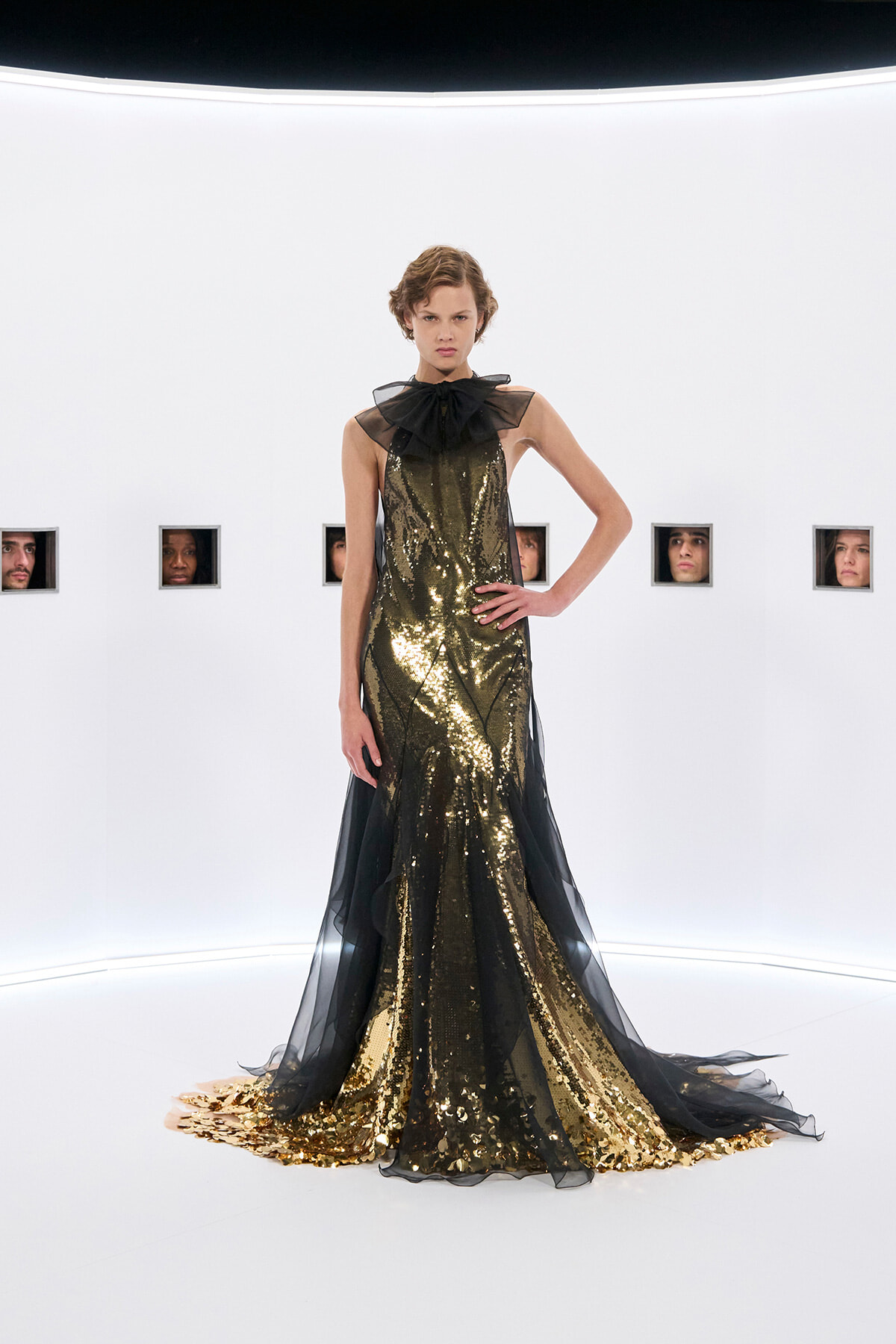 Model in a full-length gold sequin gown with black sheer overlay and a large bow at the neck, posing in a stark white studio with small square window cutouts showing faces in the background.