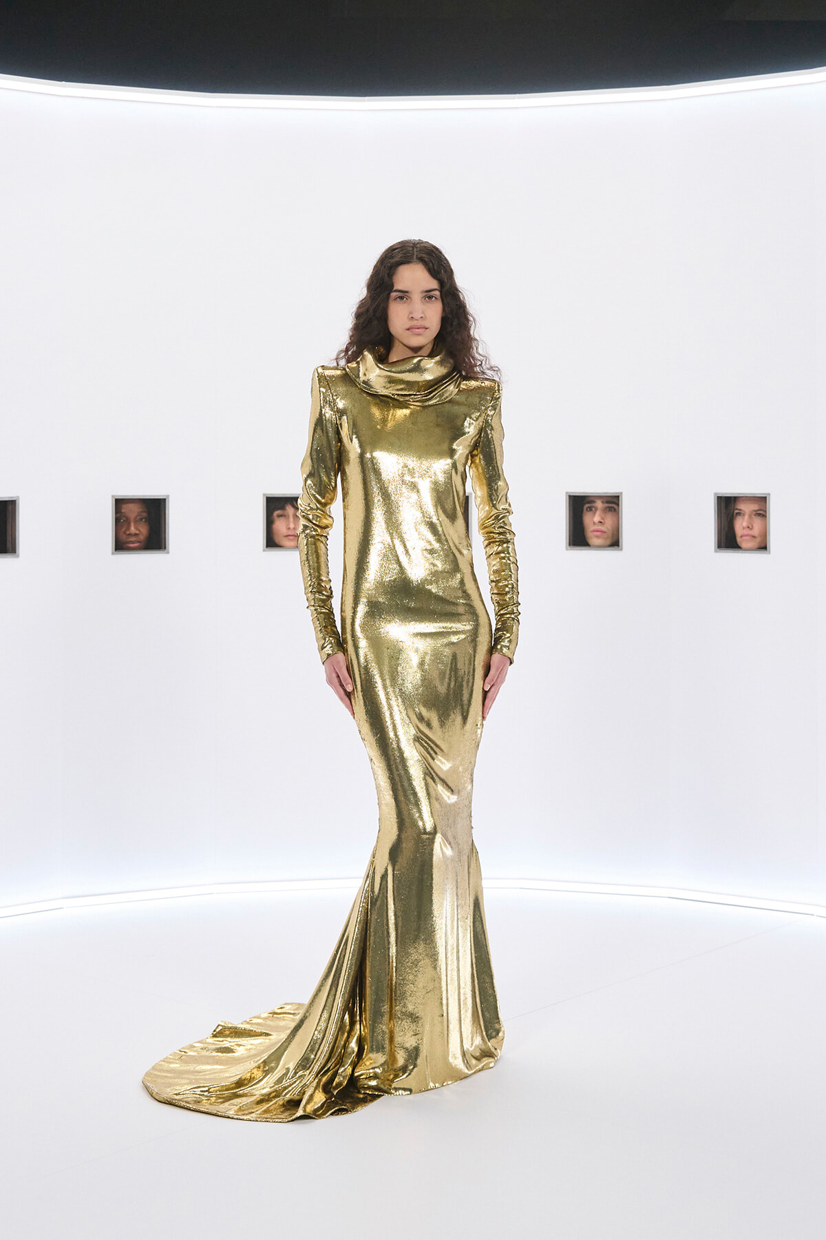 Model in a shiny gold, floor-length gown with long sleeves and a draped high neckline standing in a white studio with small square window cutouts in the wall behind her.