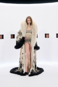 Model in a embellished gold bodysuit with a large cream fur stole and ornate cape, standing on a white studio set.