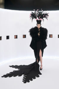 Model in a dramatic black feather cape and oversized pink-feather headdress on a white curved set, train trailing on the floor.