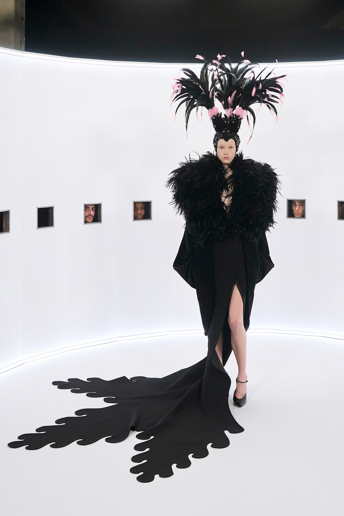 Model in a dramatic black feather cape and oversized pink-feather headdress on a white curved set, train trailing on the floor.
