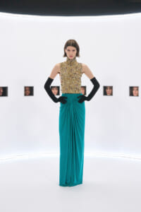 Model wearing a gold embellished high-neck top and teal draped floor-length skirt, standing with hands on hips in a bright studio; small windows with faces line the back wall.
