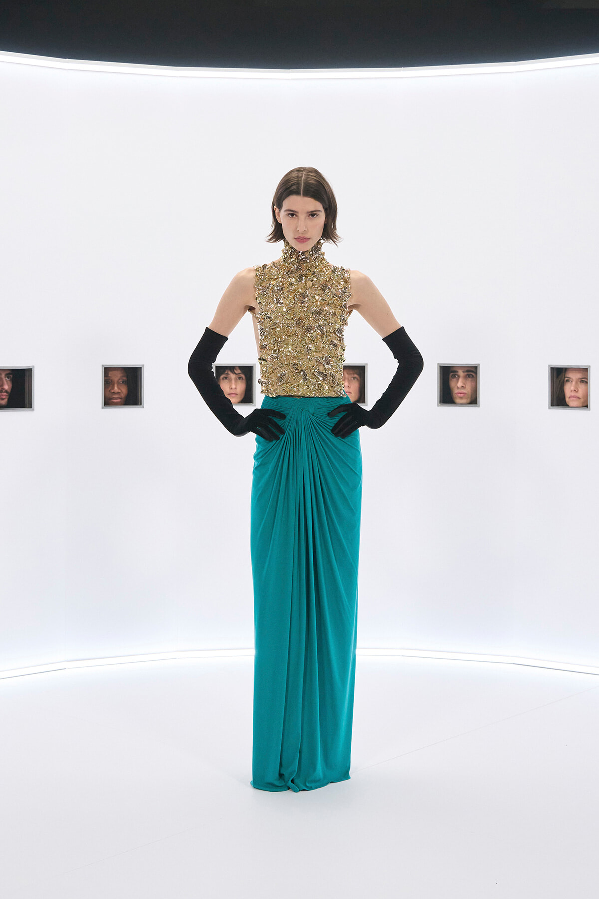 Model wearing a gold embellished high-neck top and teal draped floor-length skirt, standing with hands on hips in a bright studio; small windows with faces line the back wall.