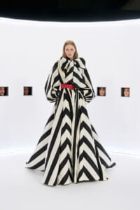 Female model wearing a black-and-white zigzag gown with an oversized bow at the neck and a red belt, posing in a stark white studio.