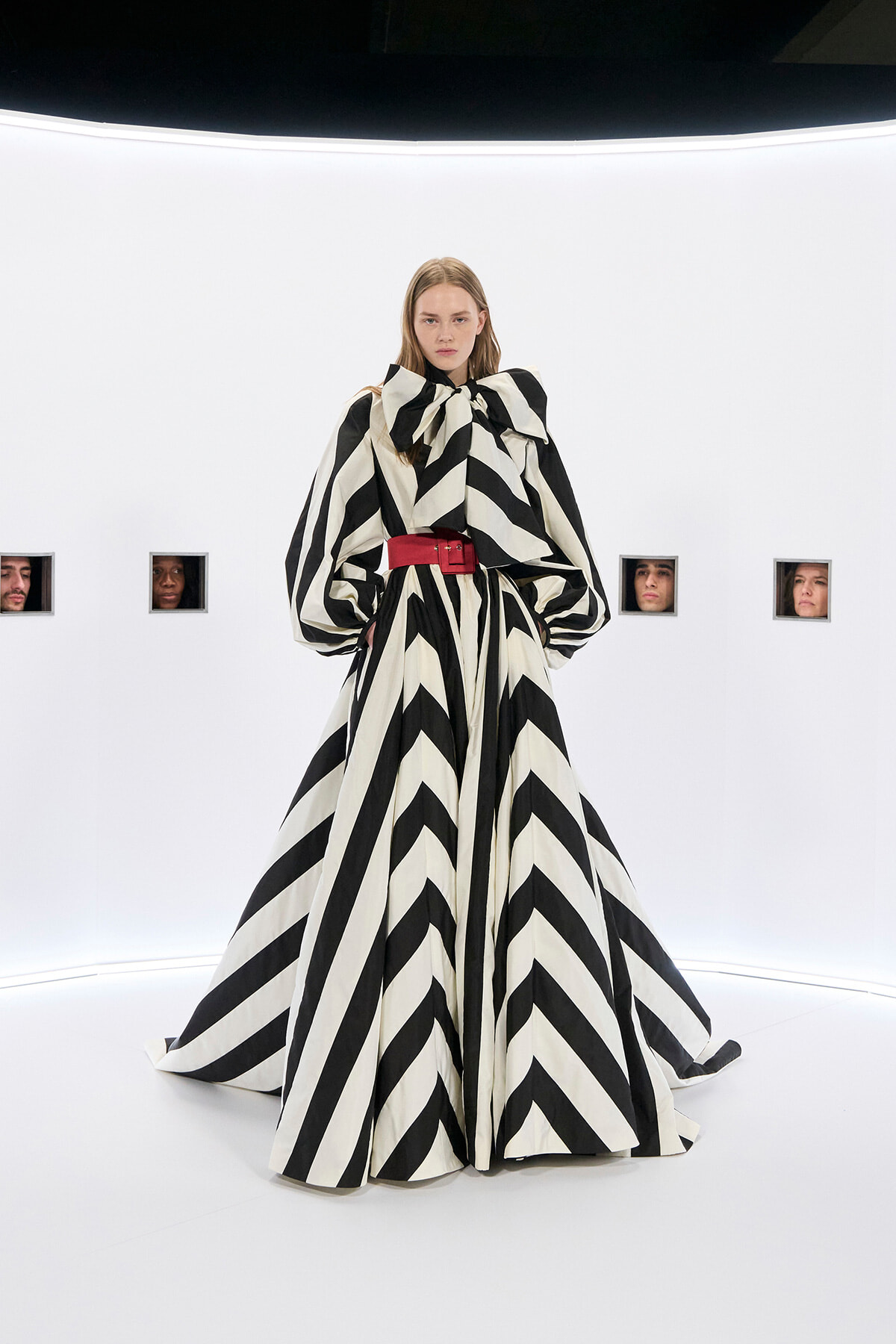 Female model wearing a black-and-white zigzag gown with an oversized bow at the neck and a red belt, posing in a stark white studio.
