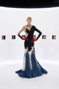 Model wearing a black-beige sequined gown with a jagged blue train and red gloves, posed in a stark white gallery setting with small portrait squares on the wall.