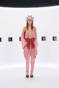 Model in a pink sequined draped jumpsuit with a large red bow, black heels, and a sunburst headpiece on a white gallery/set.