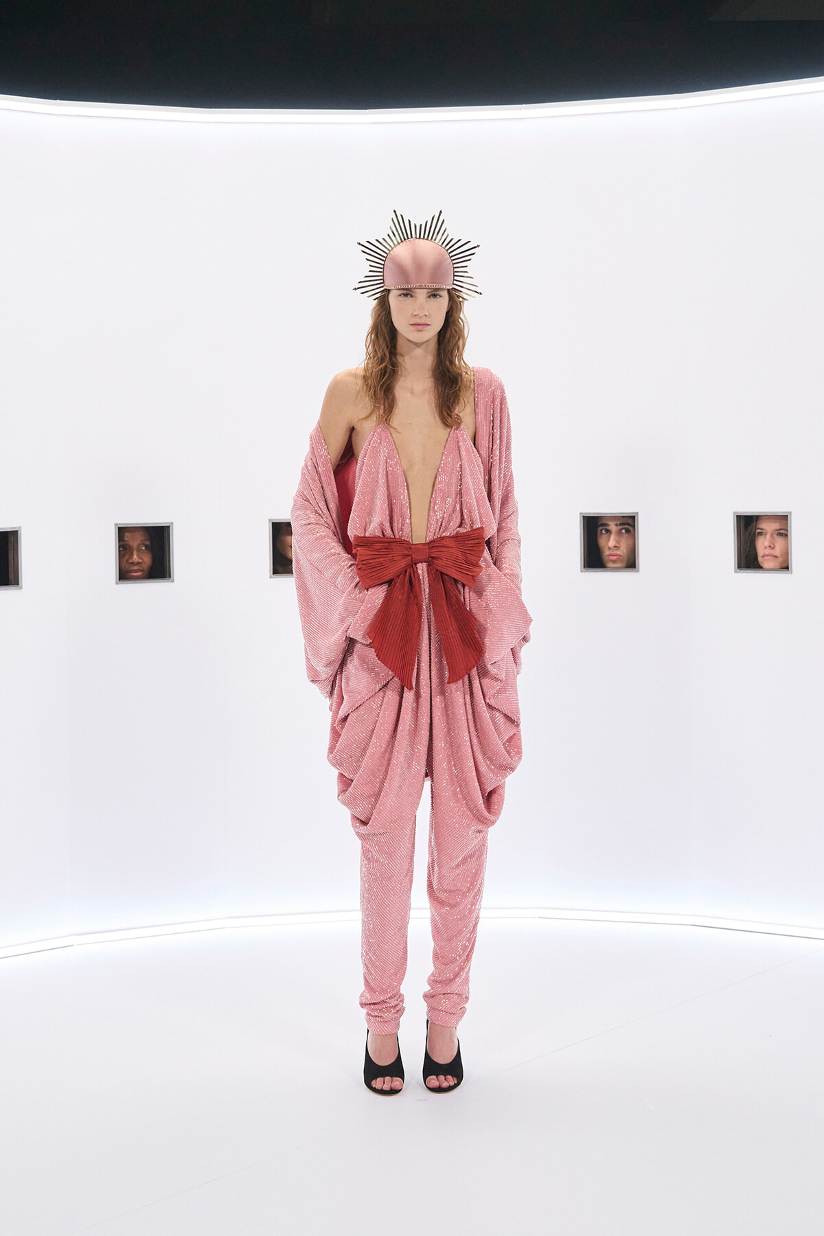 Model in a pink sequined draped jumpsuit with a large red bow, black heels, and a sunburst headpiece on a white gallery/set.