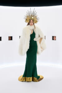 Model in an emerald green gown with a white fur cape and a gold sunburst headpiece in a bright white studio set.