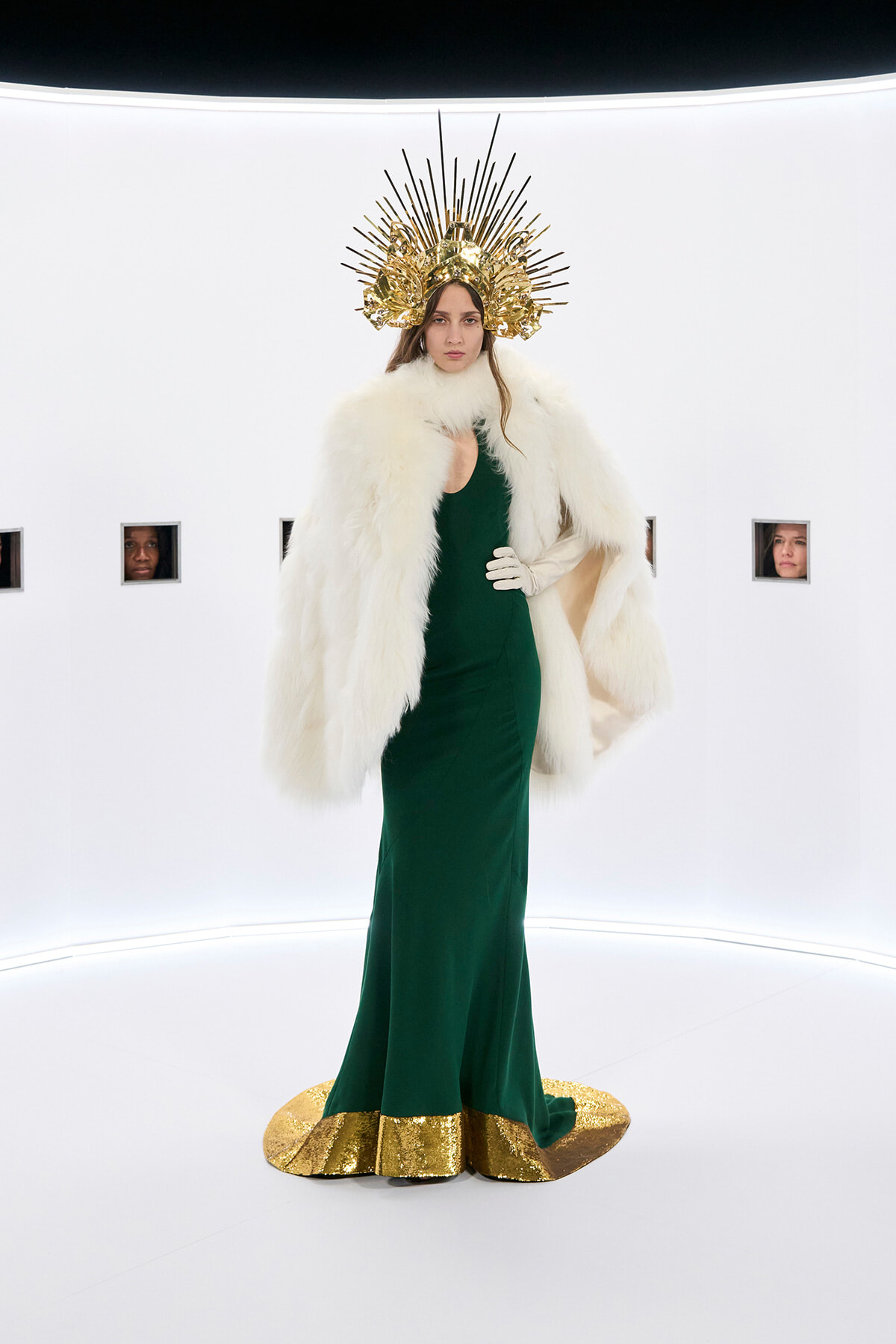 Model in an emerald green gown with a white fur cape and a gold sunburst headpiece in a bright white studio set.