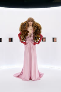 Model wearing a pale pink satin gown with an oversized gold pleated ruff and red gloves, posing in a bright white studio.