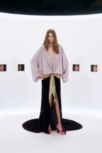 Full-body shot of a woman in a lavender, draped blouse and black skirt with a gold sash, pink heels, standing in a bright white studio. Small square faces appear in wall cutouts behind her.