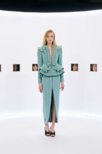 Model in aqua teal avant-garde gown with oversized shoulder ruffles and a large waist bow, standing in a white studio setting.