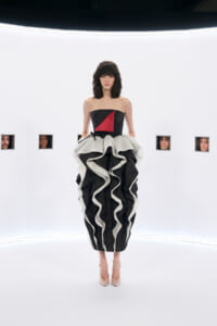 Model wearing a strapless black dress with a red geometric panel and a dramatic black-and-white ruffled skirt, standing in a stark white studio with small square portraits in the background.
