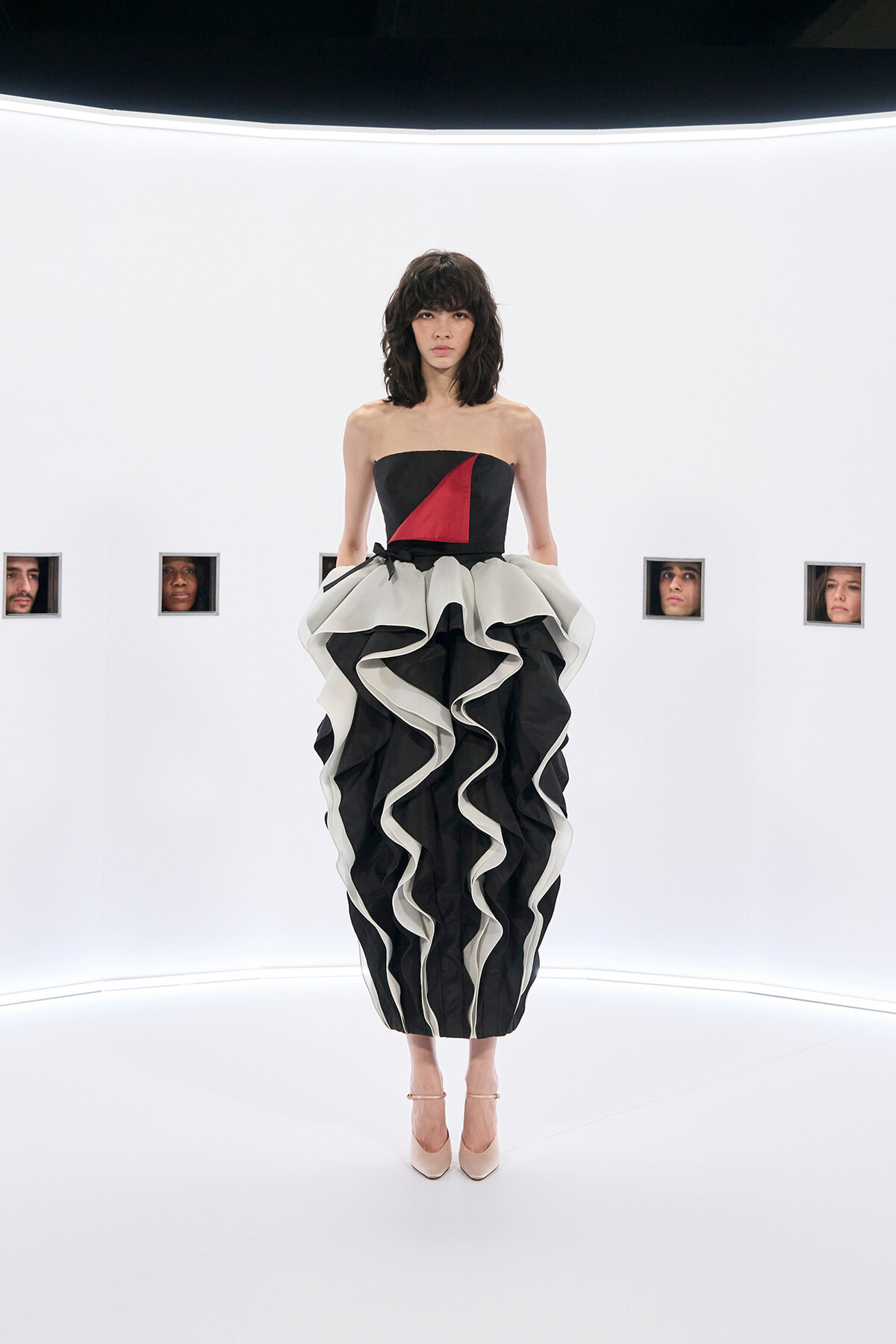 Model wearing a strapless black dress with a red geometric panel and a dramatic black-and-white ruffled skirt, standing in a stark white studio with small square portraits in the background.
