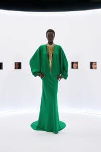 Model in an emerald green gown with dramatic voluminous sleeves and a long gold beaded necklace, standing in a stark white studio.