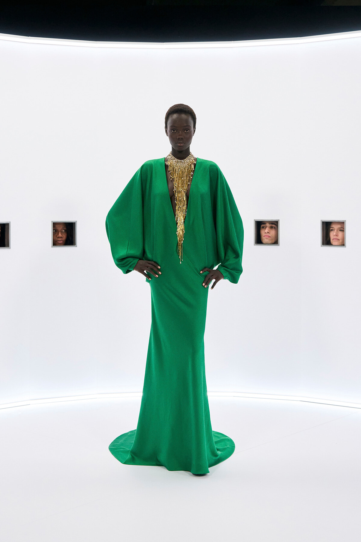 Model in an emerald green gown with dramatic voluminous sleeves and a long gold beaded necklace, standing in a stark white studio.