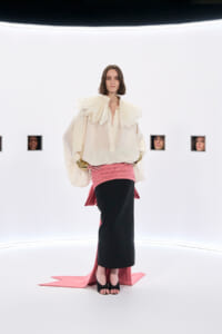 Model walks a stark white runway in a cream blouse with a dramatic pleated collar, a pink wrapped skirt over a black midi, and black open-toe heels.