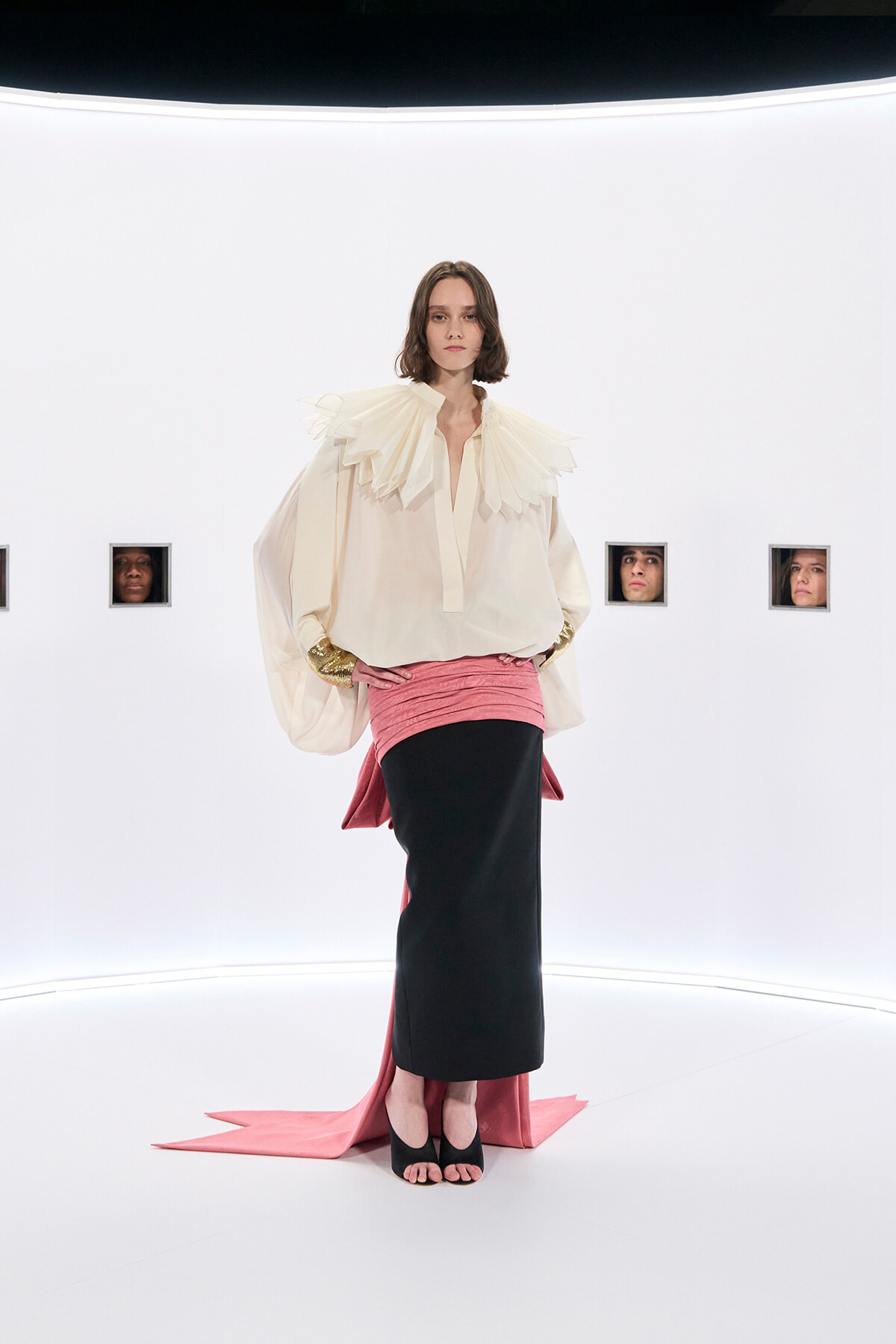 Model walks a stark white runway in a cream blouse with a dramatic pleated collar, a pink wrapped skirt over a black midi, and black open-toe heels.