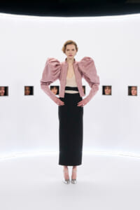 Model on a white runway wearing a pale pink structured jacket with oversized puff sleeves, a beige top, and a black ankle-length skirt with silver heels for a fashion show.