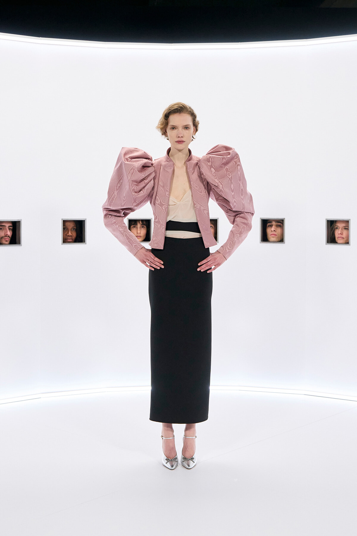 Model on a white runway wearing a pale pink structured jacket with oversized puff sleeves, a beige top, and a black ankle-length skirt with silver heels for a fashion show.