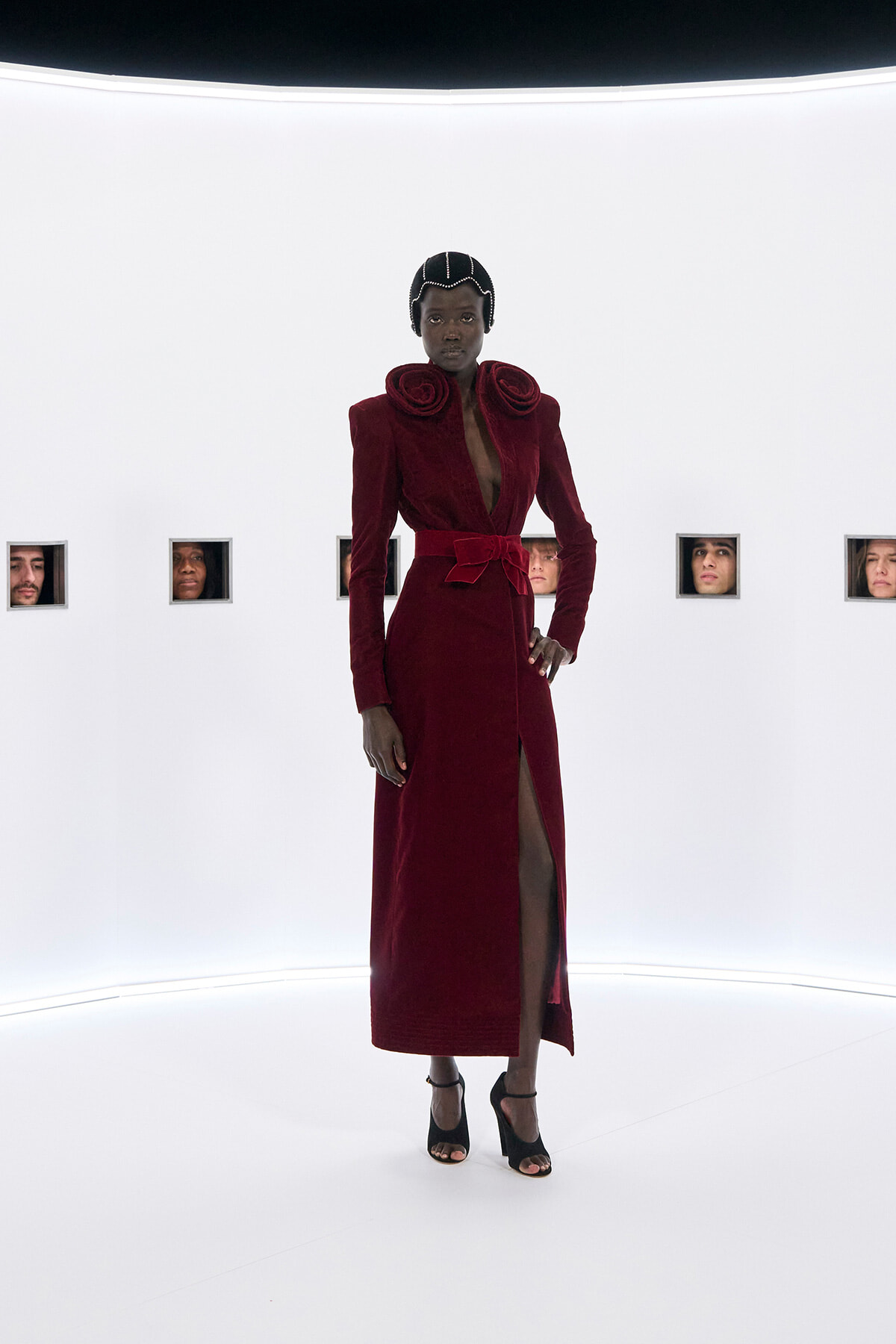 Model in burgundy velvet gown with oversized rose-shoulder details, bow belt, and high slit, posing in a white studio with square window frames showing faces