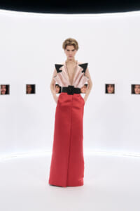 Model stands center in a pale pink top with black angular shoulder accents, a wide black belt, and a red floor-length skirt in a bright white studio background with small framed faces along the wall.