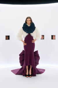 Model wearing a burgundy gown with a large teal bow and white fur stole in a stark white studio set with small square windows showing faces in the background.