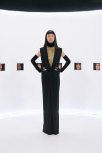 Model in a black, floor-length gown with a gold beaded bib, standing with hands on hips in a bright white studio with small square window portraits along the wall.